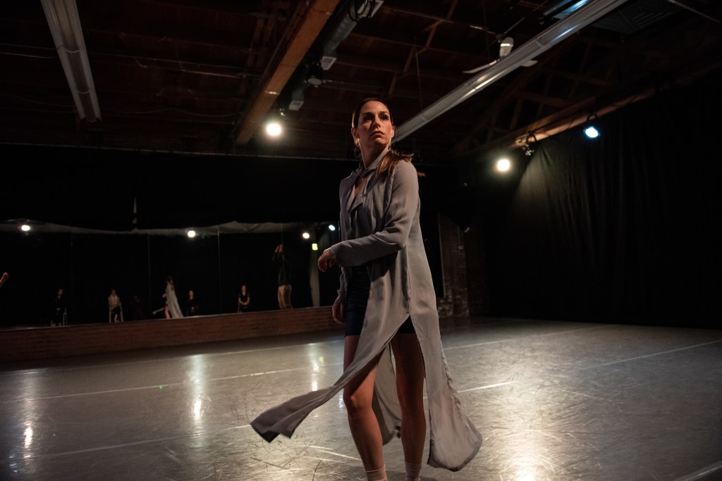 A dancer in a grey, sheer dress stares intensely at the audience, her image reflected in the mirror behind her. 