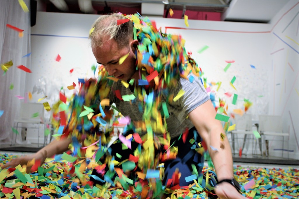 A performer is covered in confetti, they gaze downward with their head poking through; their arms are reached to the side also poking through the confetti.